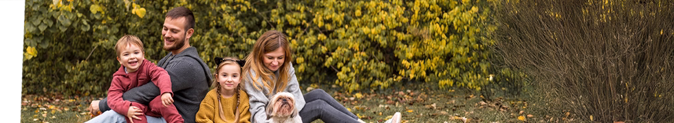 Na zdjęciu szczęśliwa rodzina odpoczywa na trawie w jesiennym parku. Tata trzyma uśmiechniętego małego chłopca w czerwonej bluzie, obok siedzi dziewczynka w żółtym swetrze, a mama przytula białego psa. Wszyscy wyglądają na zrelaksowanych i zadowolonych. W tle widoczne krzewy z żółtymi kwiatami, a wokół rozsypane jesienne liście. Obraz idealnie ilustruje wartości promowane podczas szkoleń dla rodziców w Poradni Psychologiczno-Pedagogicznej Expert, gdzie uczymy jak budować zdrowe relacje rodzinne i wspierać harmonijny rozwój dzieci.