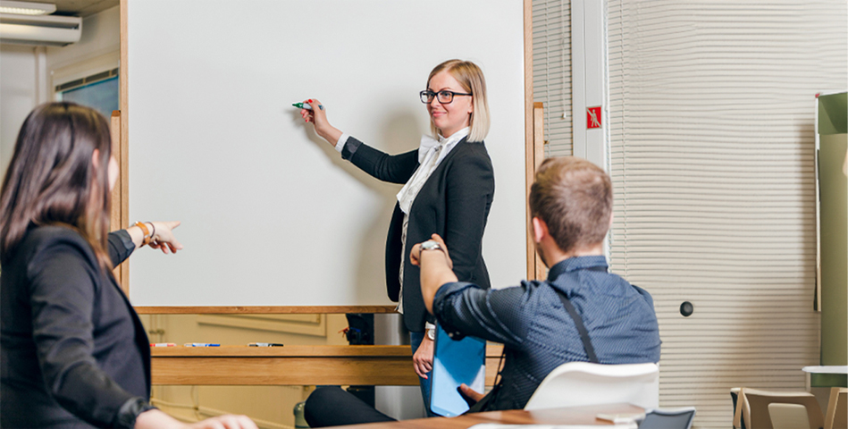 Na zdjęciu profesjonalna trenerka z rudymi włosami prowadzi szkolenie w jasnej, przestronnej sali. Ubrana w elegancką marynarkę w kratę, trzyma materiały szkoleniowe, stojąc przed tablicą flipchart. Widoczni są uczestnicy warsztatów siedzący tyłem do aparatu oraz sprzęt audio. Scena przedstawia specjalistyczne szkolenie psychologiczno-pedagogiczne, charakterystyczne dla oferty edukacyjnej Poradni Expert, gdzie doświadczeni specjaliści pomagają rozwijać kompetencje niezbędne w pokonywaniu codziennych trudności.
